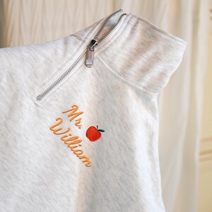 May include: A light gray sweatshirt with a silver zipper and the name "Mr. William" embroidered in orange script, along with a red apple graphic. The sweatshirt has a high collar.