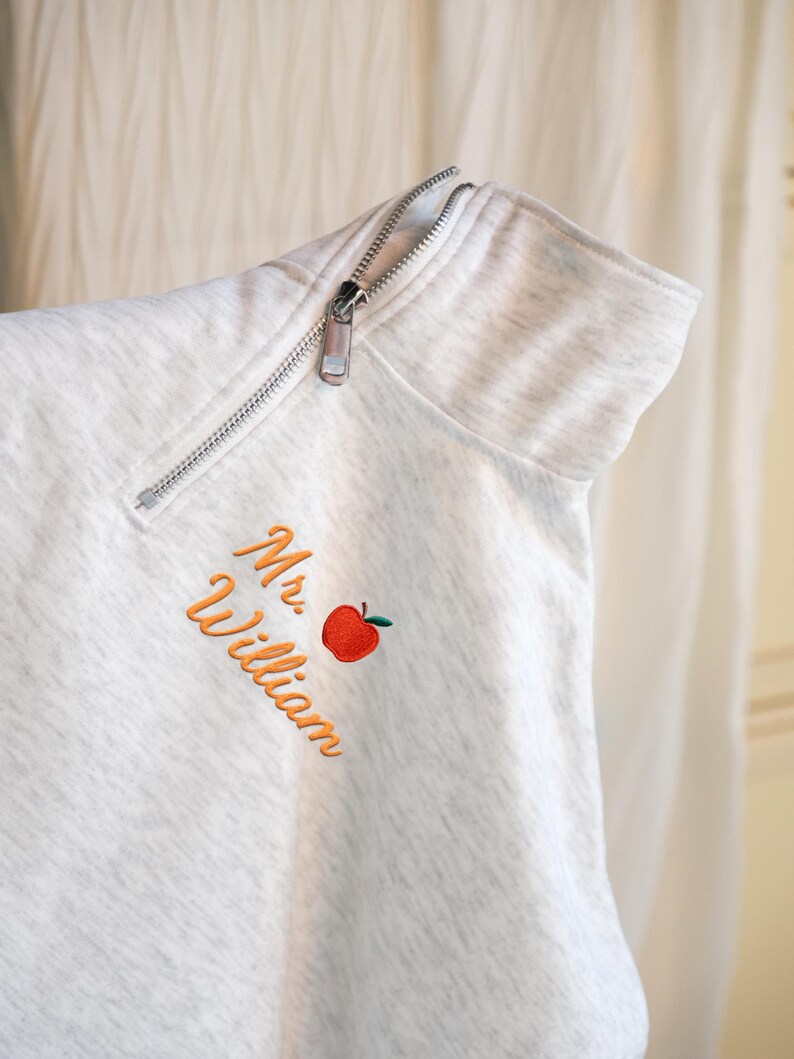 May include: A light gray sweatshirt with a silver zipper and the name "Mr. William" embroidered in orange script, along with a red apple graphic. The sweatshirt has a high collar.
