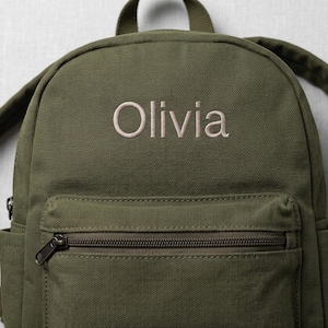 May include: An olive green canvas backpack featuring the name Olivia embroidered in a light beige thread on the upper front panel. The backpack includes a zippered front pocket, a top handle, and adjustable shoulder straps for carrying.