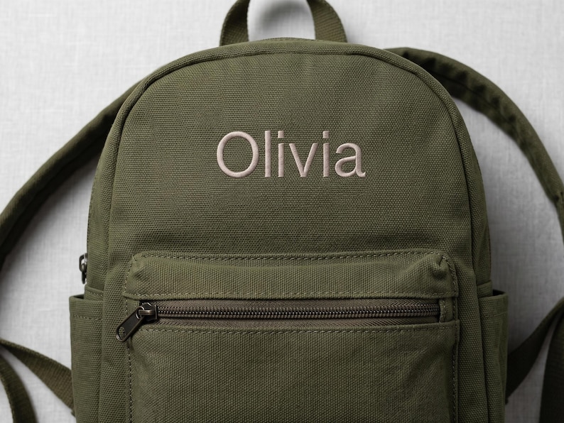 May include: An olive green canvas backpack featuring the name Olivia embroidered in a light beige thread on the upper front panel. The backpack includes a zippered front pocket, a top handle, and adjustable shoulder straps for carrying.