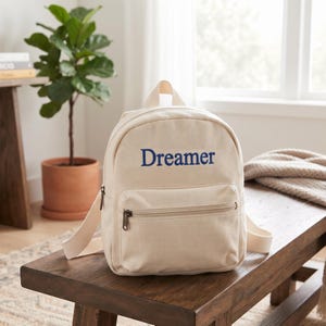 May include: A cream-colored canvas backpack featuring the word Dreamer printed in blue on the front pocket. The backpack has a top handle, adjustable shoulder straps, and a zippered front compartment with a dark metal zipper pull.