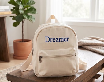 May include: A cream-coloured canvas backpack featuring the word Dreamer printed in blue on the front pocket. The backpack has a top handle, adjustable shoulder straps, and a zippered front compartment with a dark metal zipper pull.
