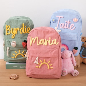 Könnte beinhalten: Drei Rucksäcke aus Cord in Grün, Rosa und Blau, jeweils mit einem Namen in einer Kontrastfarbe bestickt. Der grüne Rucksack trägt die Aufschrift "Byrdie", der rosa Rucksack "Maria" und der blaue Rucksack "Taite". Jeder Rucksack hat eine Vordertasche und ein Sonnendesign.