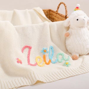 Könnte beinhalten: Cremefarbene Babydecke aus Strick mit dem Namen "Leila" in Rosa, Gelb, Orange und Blau gestickt. Ein passendes Stofflamm mit Regenbogenhut liegt daneben. Im Hintergrund ein Weidenkorb.