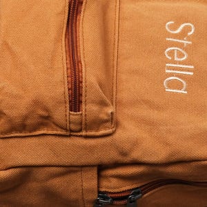 May include: A tan canvas backpack featuring a vertical zippered front pocket and the name Stella embroidered in white thread on the right side. The bag includes a main compartment with a dark brown zipper closure and matching brown zipper pulls.