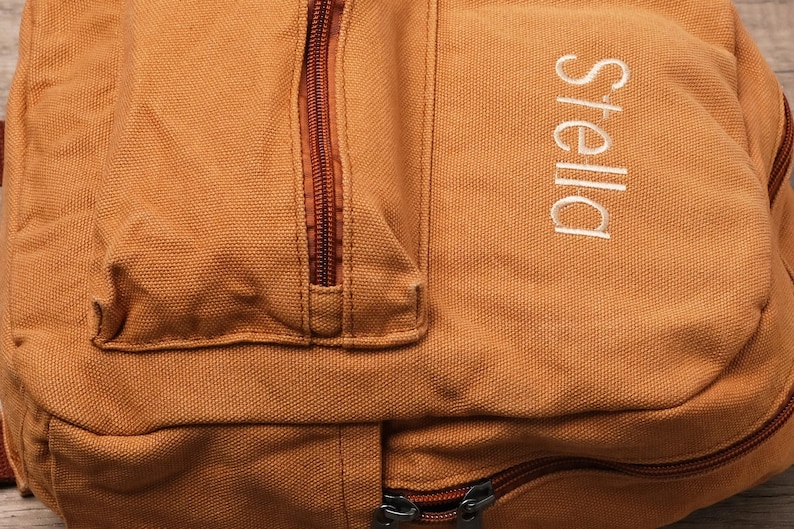 May include: A tan canvas backpack featuring a vertical zippered front pocket and the name Stella embroidered in white thread on the right side. The bag includes a main compartment with a dark brown zipper closure and matching brown zipper pulls.