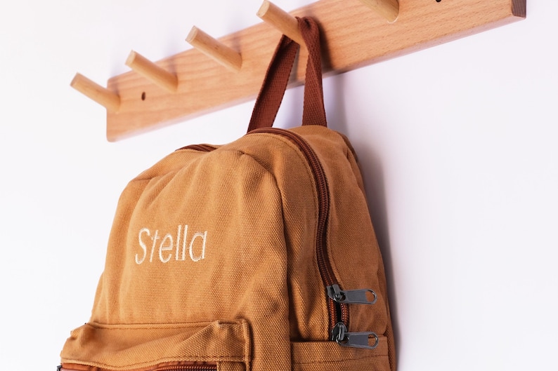 May include: A tan canvas backpack, embroidered with the name "Stella" in white, suspended from a wooden peg rail. The backpack features a brown zip and straps. The peg rail is mounted on a white wall.