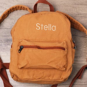 May include: A tan backpack with the name "Stella" embroidered in white. The backpack has a front pocket with a zip, brown straps, and a top handle. The background is a light wood grain.