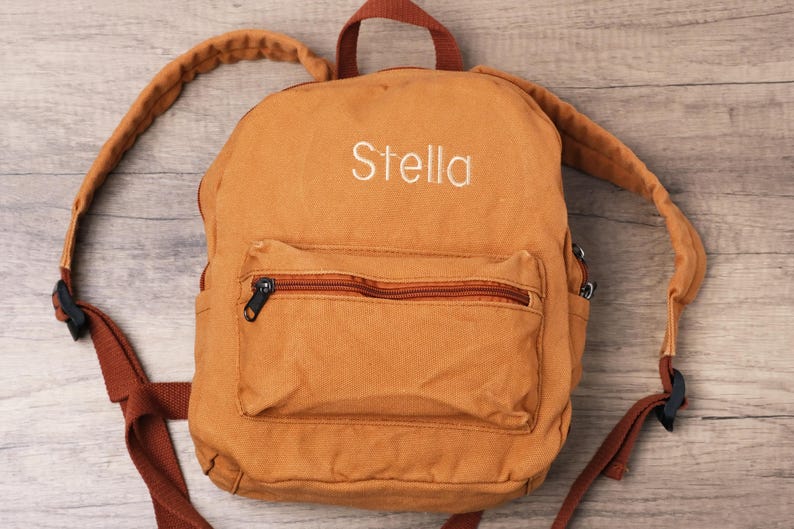 May include: A tan backpack with the name "Stella" embroidered in white. The backpack has a front pocket with a zip, brown straps, and a top handle. The background is a light wood grain.