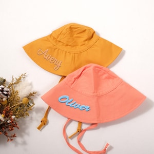 Könnte beinhalten: Zwei Sonnenhüte mit breiten Krempen. Einer ist orange mit dem Namen "Avery" in Schreibschrift gestickt. Der andere ist korallenfarben mit dem Namen "Oliver" in Blau gestickt. Beide Hüte haben Kinnriemen.