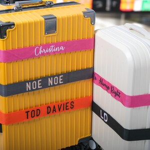Könnte beinhalten: Zwei Koffer mit Gepäckgurten. Der gelbe Koffer hat drei Gurte mit dem Text "Christina", "Noe Noe" und "Tod Davies". Der weiße Koffer hat zwei Gurte mit dem Text "Dr. Always Right" und "LD".