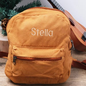 May include: Mustard-coloured backpack embroidered with the name "Stella" in white. It features a zipped front pocket and adjustable straps. A ukulele is visible in the background.