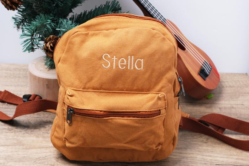 May include: Mustard-coloured backpack embroidered with the name "Stella" in white. It features a zipped front pocket and adjustable straps. A ukulele is visible in the background.