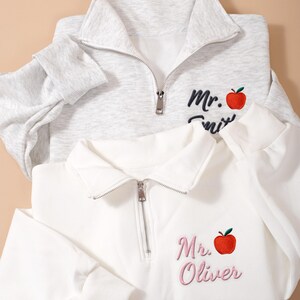 May include: Two quarter-zip pullovers, one light gray and one white. Each features an embroidered red apple and name. The gray pullover has "Mr. Smith" in black script, while the white one says "Mr. Oliver" in pink.