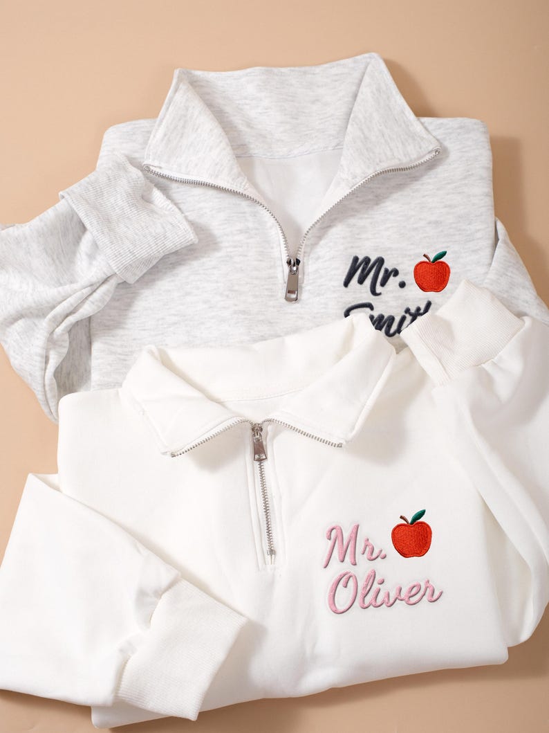 May include: Two quarter-zip pullovers, one light gray and one white. Each features an embroidered red apple and name. The gray pullover has "Mr. Smith" in black script, while the white one says "Mr. Oliver" in pink.