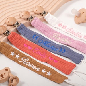 Może przedstawiać: Sześć spersonalizowanych klipsów do smoczków z różnymi wyszywanymi imionami, w tym Rowan, Emily, Marco, Ruthie, Stella i drewniany gryzak w kolorze brązowym. Klipsy są przymocowane do miękkich, kolorowych pasków z tkaniny.
