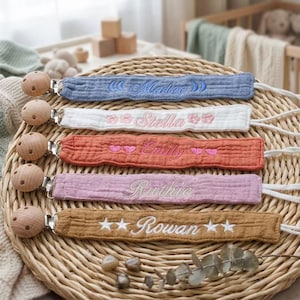 Puede incluir: Una colección de sujetachupetes personalizados para bebés, en varios colores: azul, blanco, naranja, rosa y beige. Cada uno está bordado con un nombre a juego, con detalles de cuentas de madera. Se muestran sobre una estera tejida.
