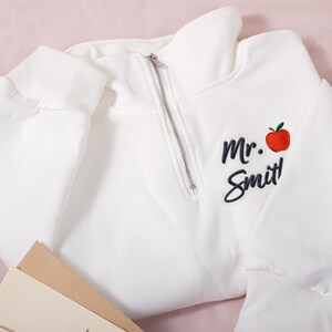 May include: White quarter-zip sweatshirt with ribbed cuffs and hem. Features the embroidered text "Mr. Smit!" with a red apple graphic. The sweatshirt is folded on a pink surface.