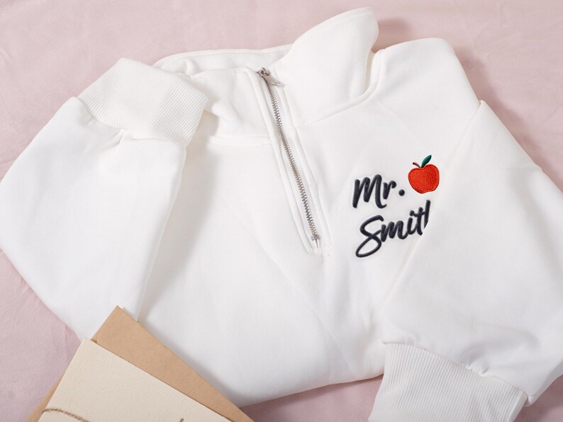 May include: White quarter-zip sweatshirt with ribbed cuffs and hem. Features the embroidered text "Mr. Smit!" with a red apple graphic. The sweatshirt is folded on a pink surface.