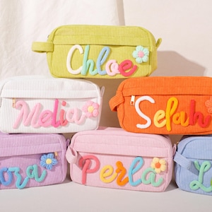 Könnte beinhalten: Eine Auswahl an Kosmetiktaschen aus Cord in verschiedenen Farben wie Grün, Weiß, Orange, Lila, Rosa und Blau. Jede Tasche ist mit einem Namen in verspielter Schrift und floralen Verzierungen versehen.