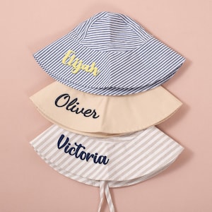 Könnte beinhalten: Drei personalisierte Sonnenhüte in einem Stapel. Der obere Hut ist blau und weiß gestreift mit dem Namen "Elijah" in gelber Schrift. Der mittlere Hut ist beige mit "Oliver" in Marineblauschrift. Der untere Hut ist gestreift mit "Victoria" in Marineblauschrift.