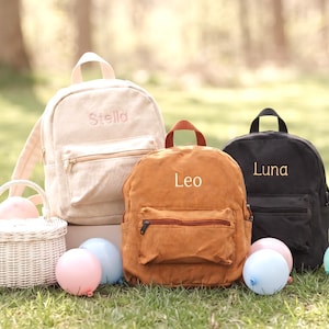 Könnte beinhalten: Drei Rucksäcke in Beige, Rostorange und Schwarz, jeweils mit einer Fronttasche mit Reißverschluss, einem Tragegriff und gestickten Namen: Stella, Leo und Luna. Daneben steht ein kleiner weißer geflochtener Korb mit Henkel und Pastellballons.