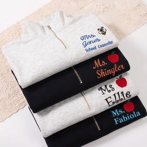 Könnte beinhalten: Vier gestapelte Pullover mit Viertelreißverschluss in Grau und Schwarz. Jeder hat einen goldenen Reißverschluss und gestickten Text. Der obere graue Pullover hat eine Biene und den Text "Mrs. Jones School Counselor". Andere Pullover haben Namen und Apfelgrafiken.