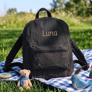 May include: A charcoal gray canvas backpack featuring a top handle, adjustable shoulder straps, and a front zippered pocket. The name Luna is embroidered in a light tan thread on the upper front section of the bag, which has a structured shape.