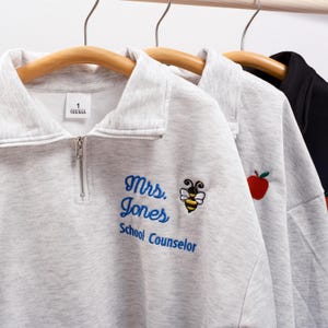 May include: Grey sweatshirt with a zip and embroidered text that reads "Mrs. Jones School Counselor" with a bee and apple design.