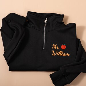 May include: Black quarter-zip sweatshirt with a high collar and a silver zipper. The sweatshirt features the name "Mr. William" embroidered in orange script with a red apple graphic. The sleeves are long and cuffed.