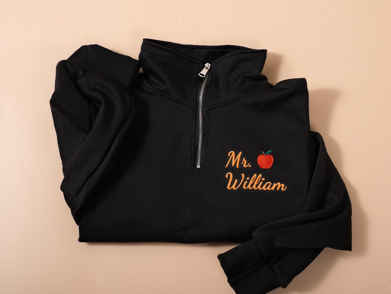 May include: Black quarter-zip sweatshirt with a high collar and a silver zipper. The sweatshirt features the name "Mr. William" embroidered in orange script with a red apple graphic. The sleeves are long and cuffed.