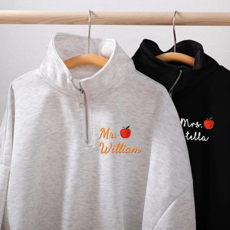 May include: Two quarter-zip sweatshirts hanging on wooden hangers. One is light gray with "Mr. William" and an apple graphic embroidered in orange. The other is black with "Mrs. Stella" and an apple graphic.