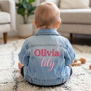Könnte beinhalten: Hellblaue Jeansjacke mit dem Namen "Olivia lily" in Rosa auf dem Rücken gestickt. Die Jacke ist geknöpft und hat einen klassischen Kragen. Das Kind sitzt auf einem weiß-beigen Teppich.