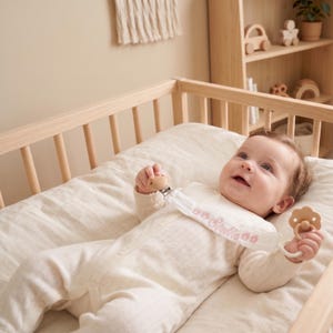 May include: A baby in a cream-colored onesie lies in a wooden crib, holding a wooden pacifier and a matching pacifier clip. The clip is white with the name "Stella" embroidered in pink. The crib is in a light-toned room.
