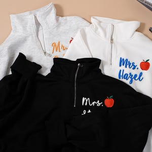 May include: Three quarter-zip sweatshirts in gray, white, and black. Each sweatshirt has the name "Mrs." embroidered on it, along with a red apple graphic. The white sweatshirt has "Mrs. Hazel" embroidered in blue. The gray sweatshirt has "Mrs." embroidered in orange.