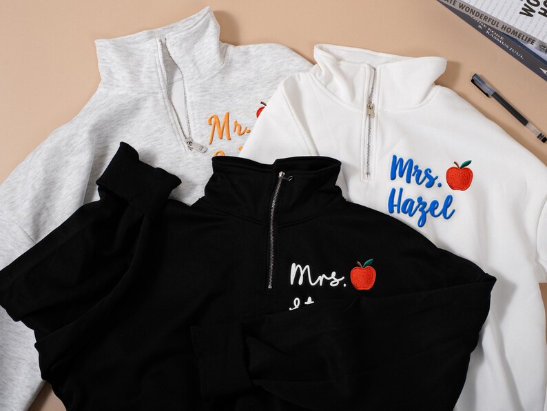 May include: Three quarter-zip sweatshirts in gray, white, and black. Each sweatshirt has the name "Mrs." embroidered on it, along with a red apple graphic. The white sweatshirt has "Mrs. Hazel" embroidered in blue. The gray sweatshirt has "Mrs." embroidered in orange.