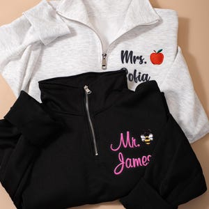May include: Two quarter-zip pullovers, one light gray and one black. The gray pullover has "Mrs. Sofia" embroidered with a red apple. The black pullover has "Mr. James" embroidered with a bee. Both have ribbed cuffs.