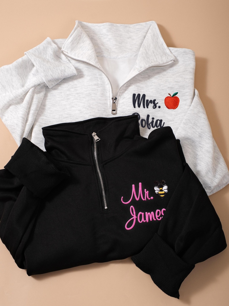 May include: Two quarter-zip pullovers, one light gray and one black. The gray pullover has "Mrs. Sofia" embroidered with a red apple. The black pullover has "Mr. James" embroidered with a bee. Both have ribbed cuffs.