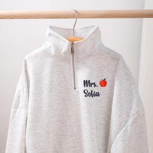 Könnte beinhalten: Ein hellgraues Sweatshirt mit Stehkragen und einem Viertel-Reißverschluss. Das Sweatshirt hat den Text "Mrs. Sofia" mit einer roten Apfelgrafik auf der Vorderseite. Das Sweatshirt hängt an einer Holzstange.