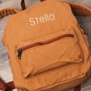 May include: A tan canvas backpack with the name "Stella" embroidered in white. The backpack has a front pocket with a brown zip and black zip pull. Adjustable brown straps and black buckles are visible.