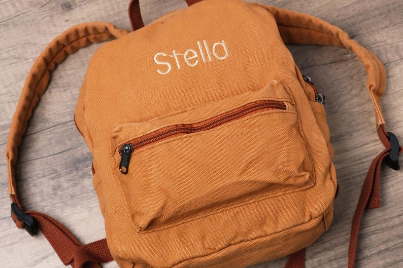 May include: A tan canvas backpack with the name "Stella" embroidered in white. The backpack has a front pocket with a brown zip and black zip pull. Adjustable brown straps and black buckles are visible.