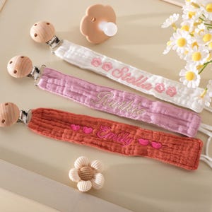 Könnte beinhalten: Drei Baby-Schnullerclips in Weiß, Rosa und Orange mit gestickten Namen und Blumen- oder Herzmotiven. Ein beiger Schnuller und eine dekorative Blume sind ebenfalls vorhanden. Die Clips haben Holzakzente.
