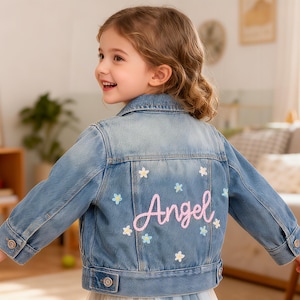 Könnte beinhalten: Hellblaue Jeansjacke mit dem Namen "Angel" in rosa Schrift auf dem Rücken gestickt. Die Jacke ist mit kleinen gestickten Blumen in Weiß, Hellblau und Rosa verziert. Die Jacke hat Knopfverschlüsse und Taschen.