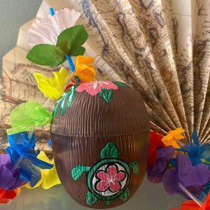 May include: A brown coconut-shaped cup with a painted green sea turtle and pink flower design. The cup is surrounded by colorful leis and a paper fan with a map design.