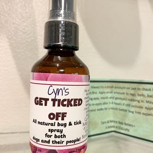 May include: A brown glass spray bottle with a black spray nozzle. The label reads "Cyn's GET TICKED OFF" and advertises an all-natural bug and tick spray for dogs and people. The label has a pink floral design.