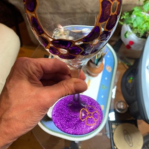 May include: A purple wine glass with a gold rim and purple flowers painted on the glass. The glass is decorated with glitter.