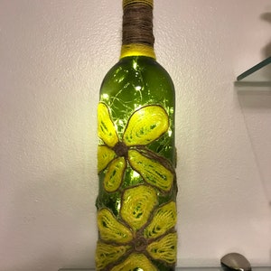 May include: A green glass bottle with a cork top decorated with yellow yarn flowers. The bottle is lit from inside with white fairy lights.