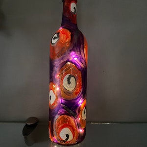 May include: A purple glass bottle with a cork top is decorated with orange and white painted eyes. The bottle is lit from inside with white fairy lights.
