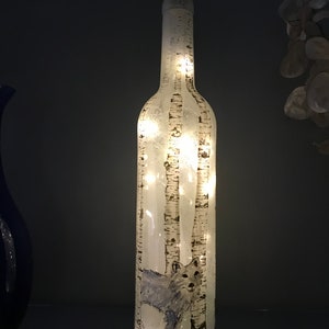 May include: A clear glass bottle with a white wolf silhouette and birch tree design. The bottle is lit from within with white fairy lights.