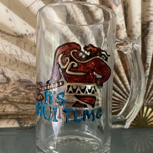 Puede incluir: Una jarra de cerveza de vidrio transparente con un dise&ntilde;o pintado a mano de Maui de la pel&iacute;cula Moana. El dise&ntilde;o est&aacute; en tonos de marr&oacute;n, dorado y azul. El texto "It's Maui Time" est&aacute; escrito en azul en la jarra.